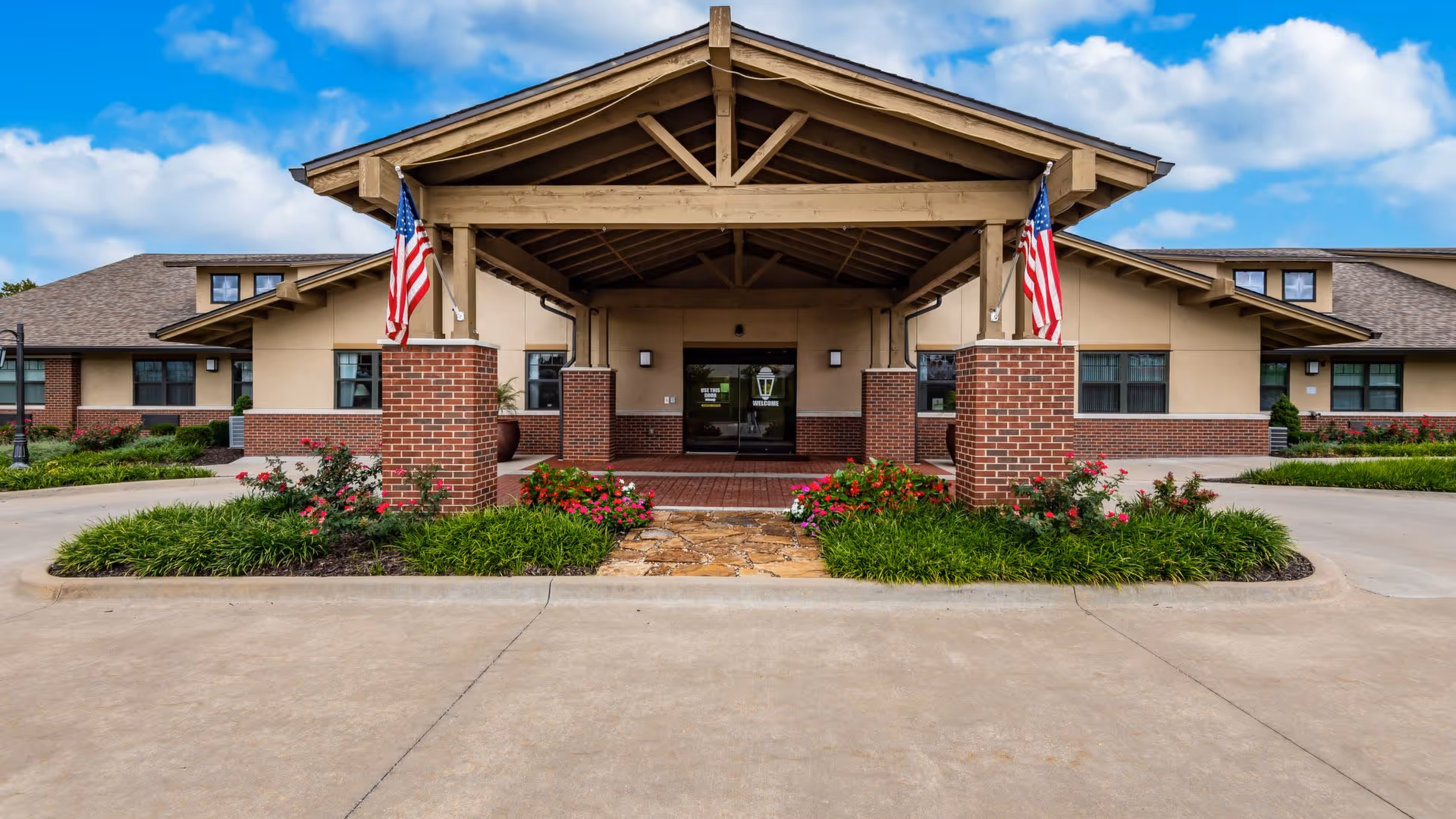 Front entrance of Morningside Place facility featuring a covered porch with wooden beams and brick pillars, two American flags, landscaped flower beds with green plants and red flowers, and a clear blue sky with some clouds.