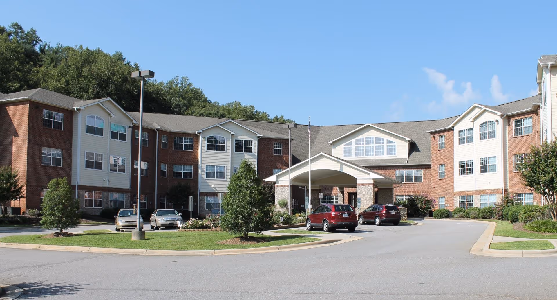 Front exterior view of Rose Glen Village, a multi-story senior living facility with a covered entrance, several parked cars, and well-maintained landscaping under a clear blue sky.