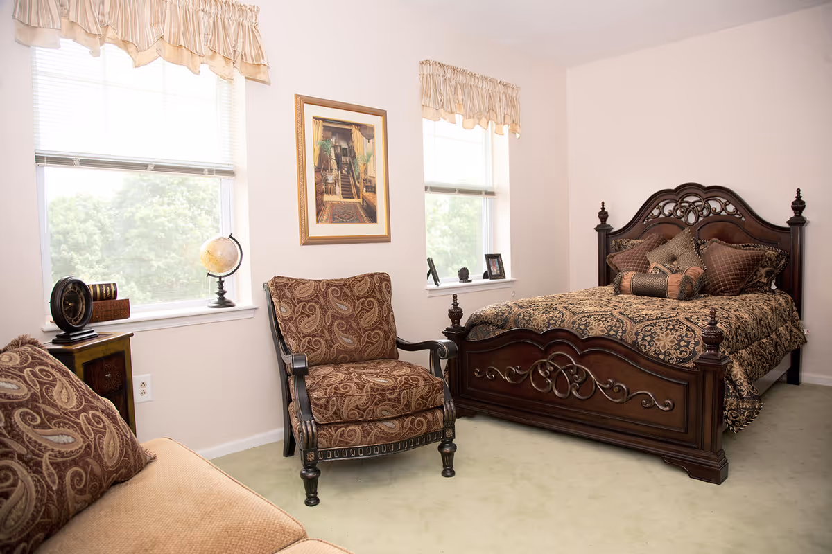 A bedroom with a dark wooden bed frame featuring intricate carvings, a matching armchair with patterned upholstery, a beige sofa with patterned cushions, two windows with blinds and valances, a small wooden side table with a globe and decorative items, and a framed picture on the wall between the windows.