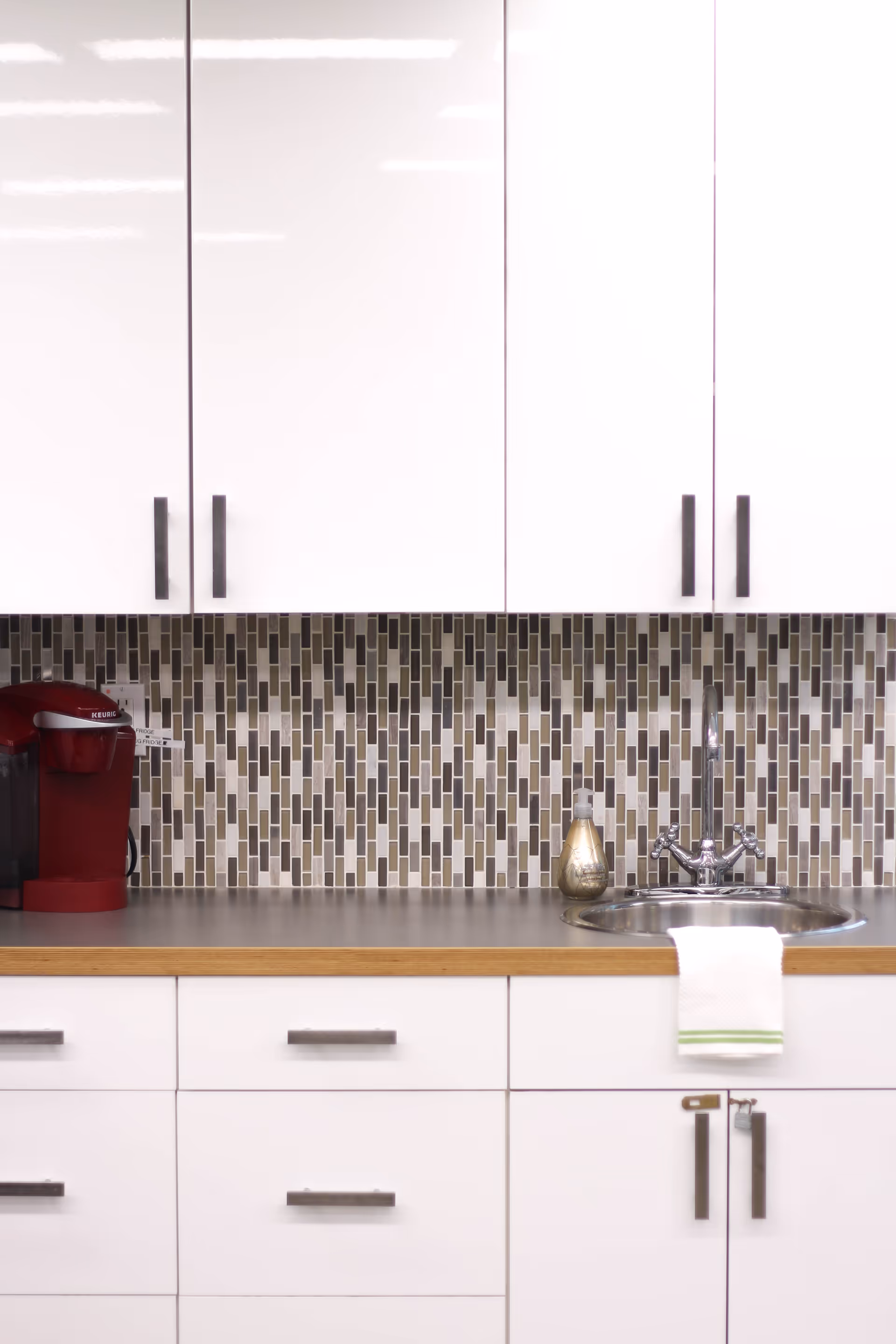 A modern kitchen area with white cabinets and drawers, a countertop with a built-in round sink and a chrome faucet. There is a red Keurig coffee maker on the left side of the counter and a soap dispenser next to the sink. A white towel with a green stripe hangs over the edge of the counter. The backsplash features a mosaic tile pattern in shades of brown, beige, and white.