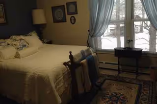 Cozy bedroom with a bed, nightstand, lamp, window with blue curtains, and rug.