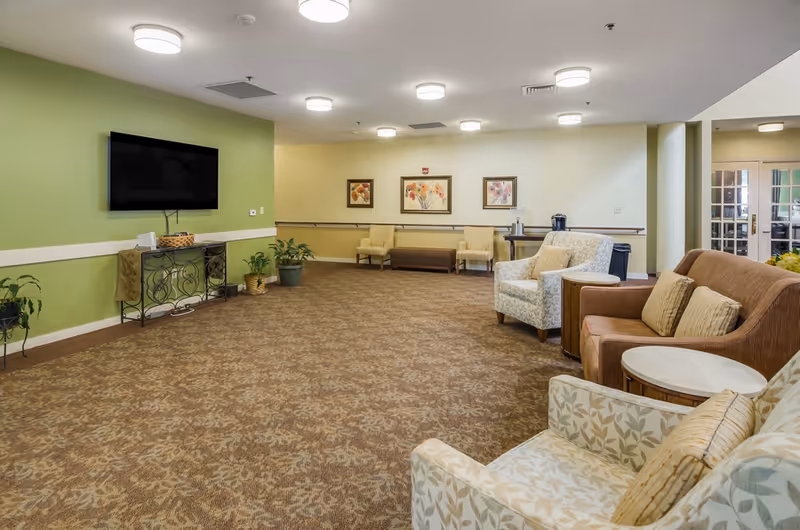 A spacious living room area in a senior living facility with patterned carpet, several upholstered chairs and sofas arranged around small round tables, a flat-screen TV mounted on a green accent wall, potted plants, and framed floral artwork on the walls.
