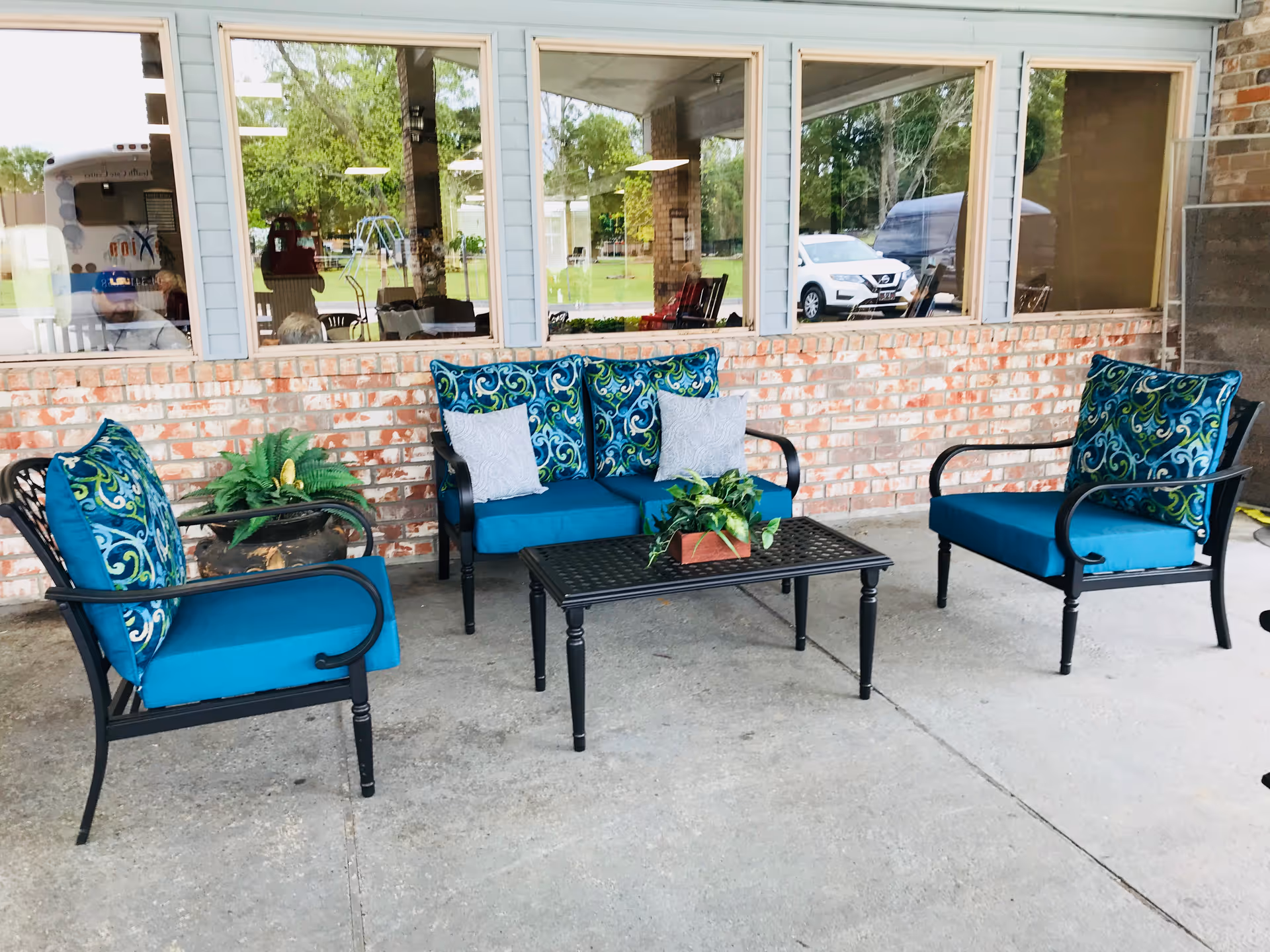 Covered outdoor patio area with teal-cushioned chairs and a loveseat around a black coffee table in front of a brick wall and windows.