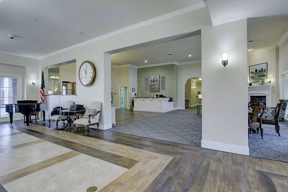 Spacious and well-lit senior living facility common area with a grand piano near a window, two armchairs with a small table between them, a large wall clock, and a reception desk in the background. The room features a mix of wood and carpet flooring, wall sconces for lighting, and decorative artwork on the walls.