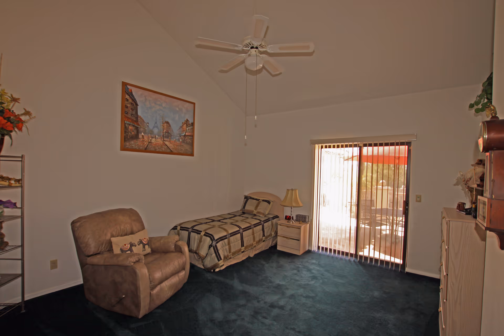 A bedroom with a single bed covered in a plaid blanket, a beige recliner chair with a decorative pillow, a nightstand with a lamp and alarm clock, a dresser, and a sliding glass door with vertical blinds leading to an outdoor patio. The room has a ceiling fan and a framed painting on the wall.