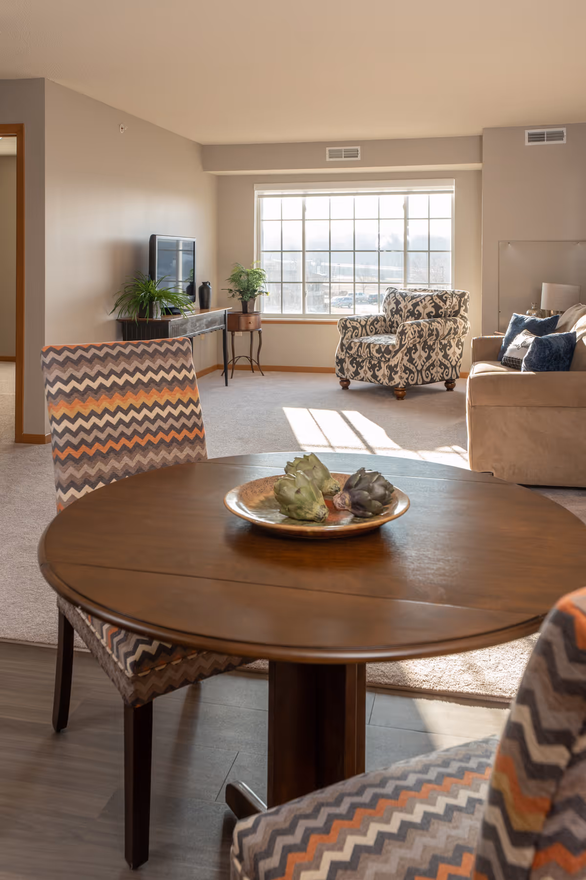 A bright living room area with a large window letting in natural light. In the foreground, there is a round wooden table with a decorative plate holding artichokes. Two chairs with zigzag patterned upholstery surround the table. In the background, there is a patterned armchair, a beige sofa with blue cushions, a small table with plants, and a TV on a stand against the wall.