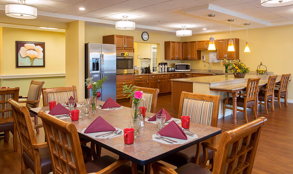 A dining area in a senior living facility featuring a table set with red napkins, red mugs, glassware, and silverware. The room has wooden chairs with cushioned seats, a kitchen area with wooden cabinets, a stainless steel refrigerator, an oven, and a countertop with bar stools. The walls are painted yellow, and there is a floral painting on one wall. Pendant lights hang over the kitchen counter.
