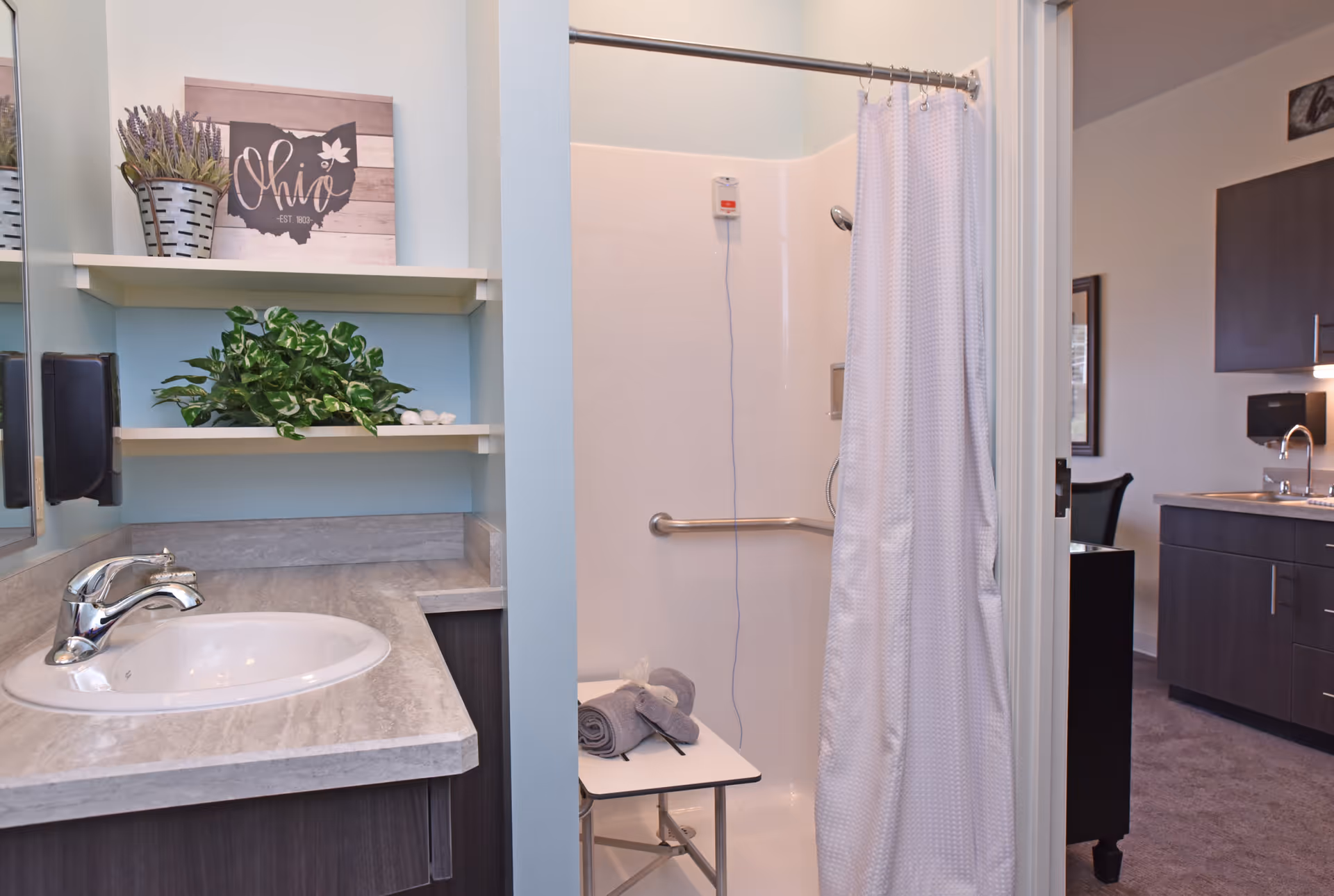 A bathroom with a sink and countertop on the left, two shelves with decorative plants and a wooden Ohio sign above the sink, and a shower with a white curtain and a foldable shower seat inside. The bathroom opens into a room with dark cabinetry and a sink visible in the background.