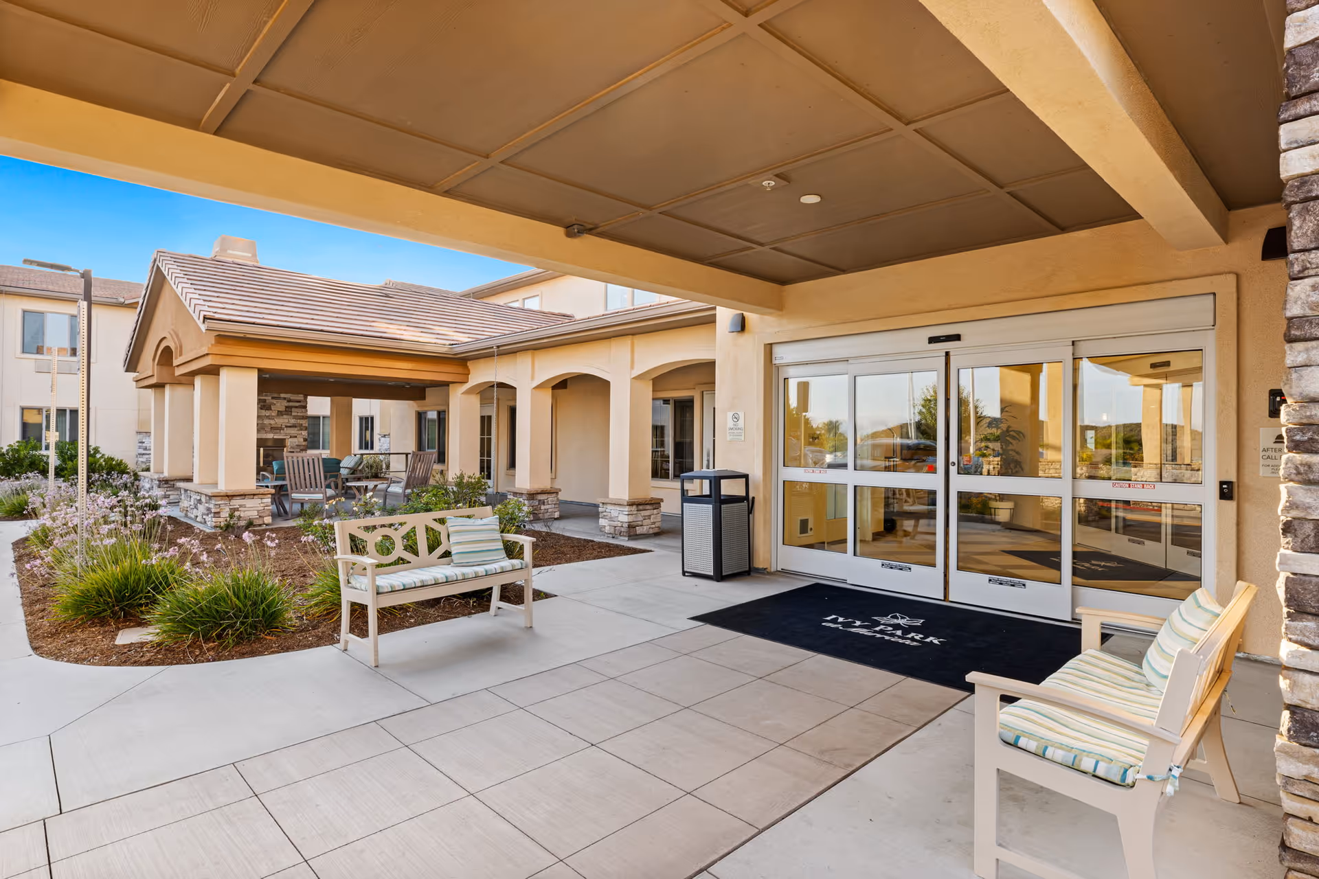 Entrance area of Ivy Park At Murrieta facility showing a covered walkway with cushioned benches, a black welcome mat with the facility's name, glass automatic doors, and a landscaped garden area with outdoor seating under a pergola.
