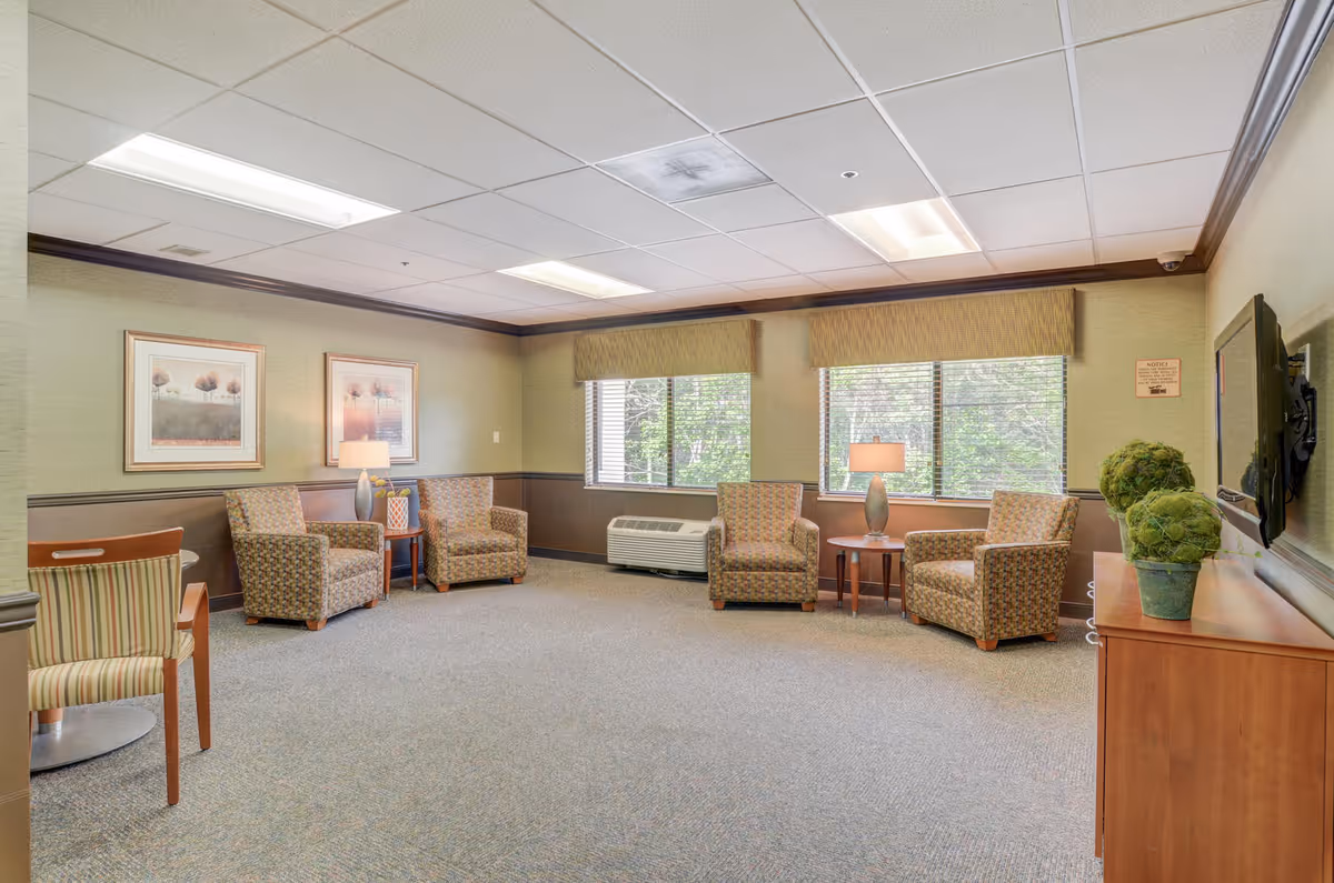 A bright communal sitting room with upholstered armchairs, side tables and lamps, large windows, and a wall-mounted TV.