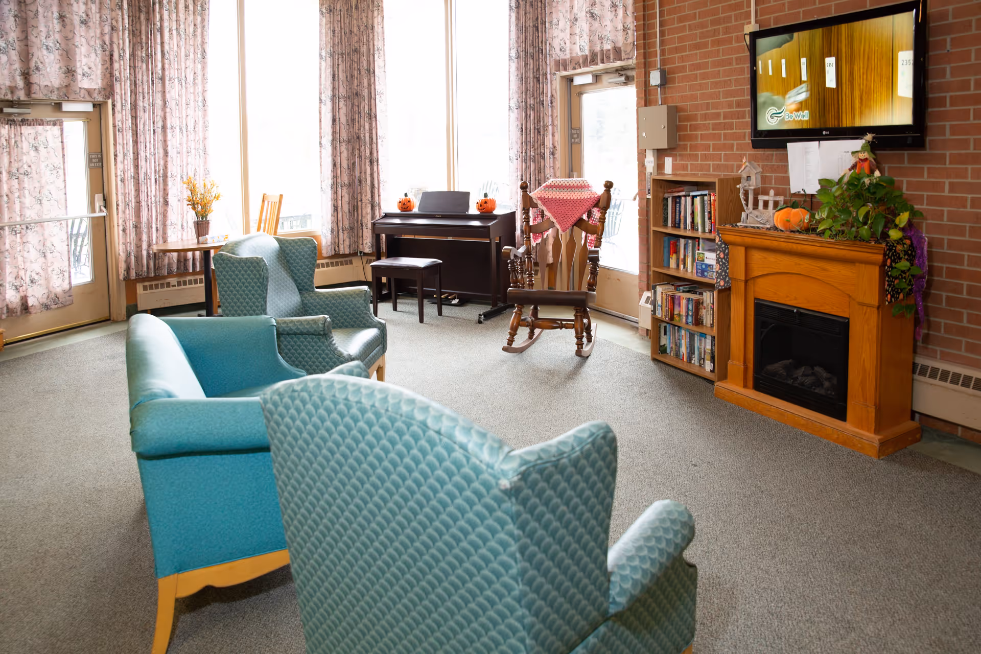A cozy living room with teal upholstered chairs and a sofa arranged around a wooden electric fireplace. A rocking chair with a pink crocheted blanket sits near a small bookshelf filled with books. A piano with two small pumpkin decorations is placed against the window with floral curtains. A flat-screen TV is mounted on a brick wall above the fireplace, and a small table with a vase of yellow flowers is near a glass door.