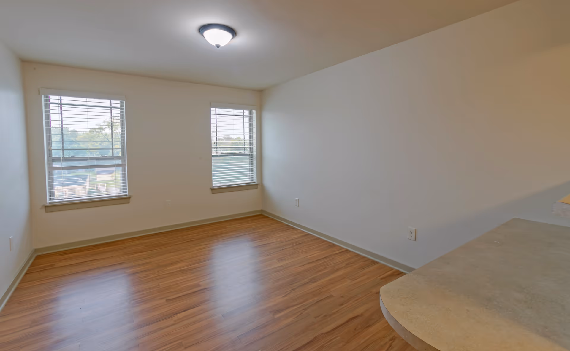Empty bright room with wood floors, two windows with blinds and a small countertop visible in the foreground.