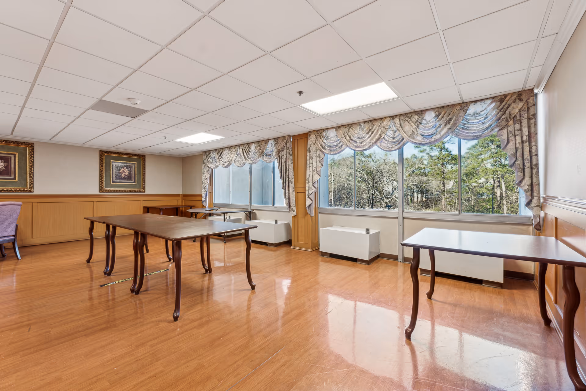 Sunlit dining/activity room with wooden tables, large windows with patterned valances, framed artwork, and polished wood floors.