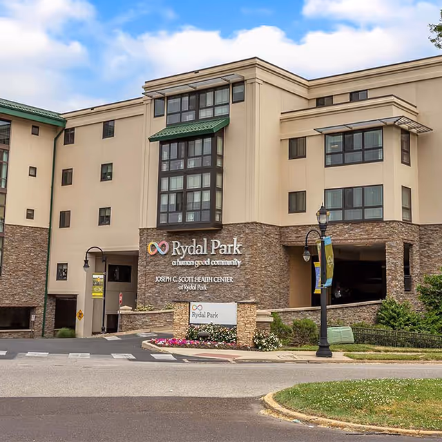 Exterior view of the Rydal Park building showing a multi-story structure with beige walls and stone accents. The building has several windows, green awnings, and a sign that reads 'Rydal Park a human good community JOSEPH C. SCOTT HEALTH CENTER of Rydal Park'. There is a street with a crosswalk and a lamp post with banners in front of the building.