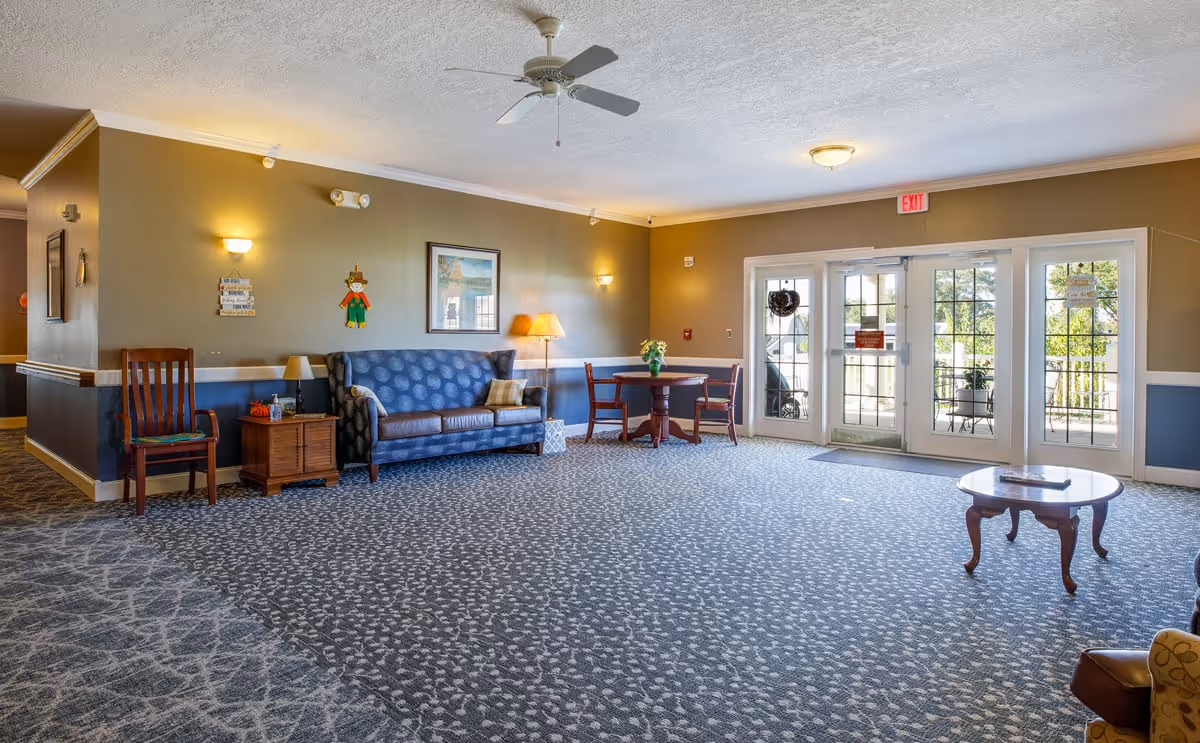 A spacious and well-lit common area in an assisted living facility featuring a blue patterned sofa with pillows, wooden chairs, a small round table with a flower vase, a wooden side table with a lamp, and large glass doors leading to an outdoor patio. The room has beige and blue walls, patterned carpet flooring, and ceiling lights with a ceiling fan.
