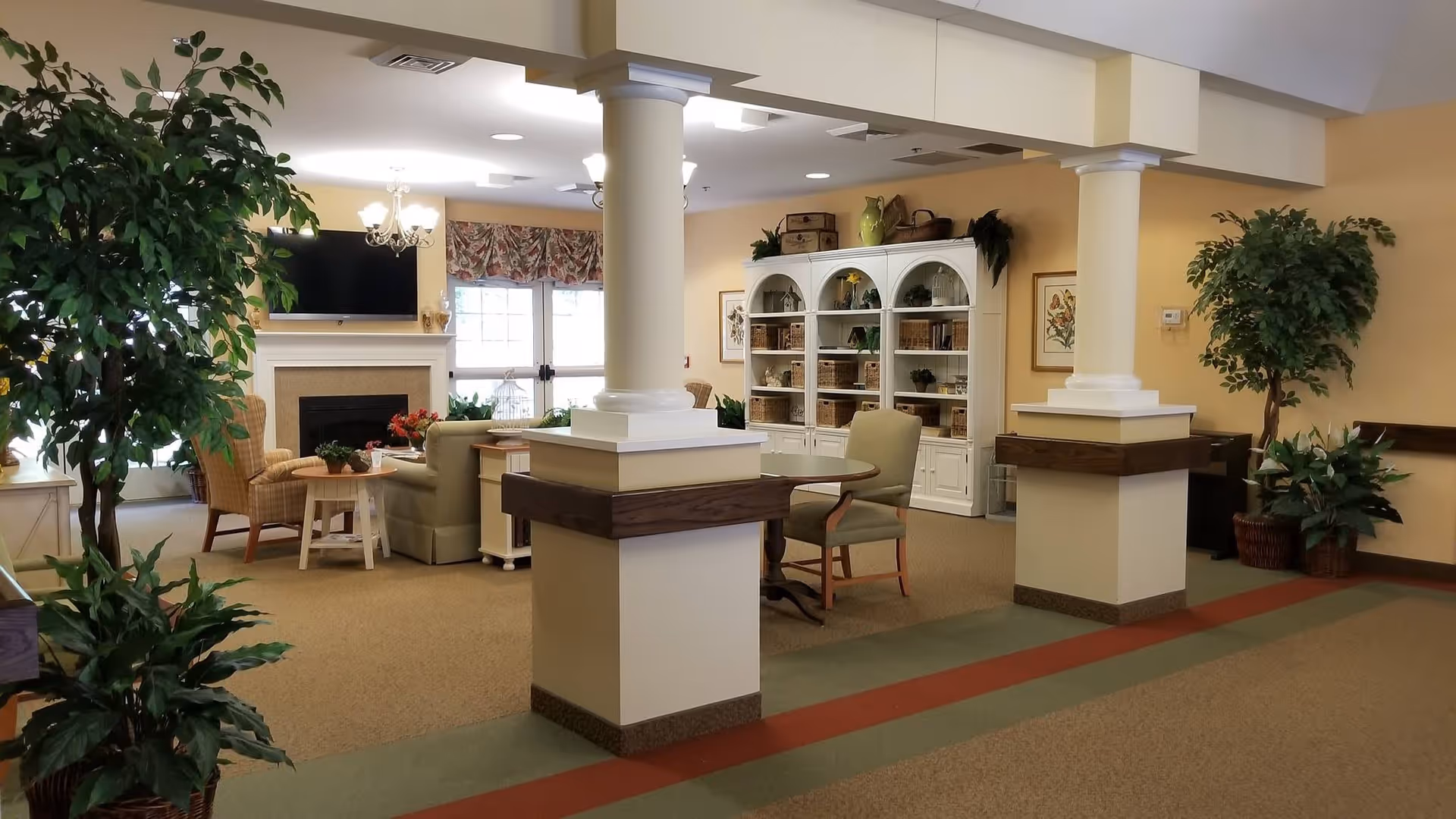 Bright communal living room with chairs and tables arranged around a fireplace and TV, white bookshelves, plants, and decorative columns.