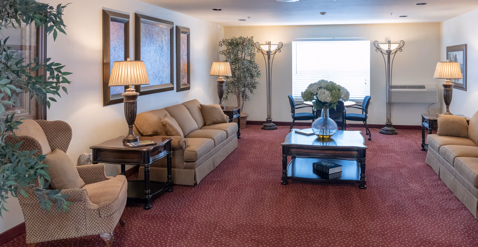 Well-lit communal living room with sofas, armchairs, a central coffee table, floor and table lamps, and decorative plants.