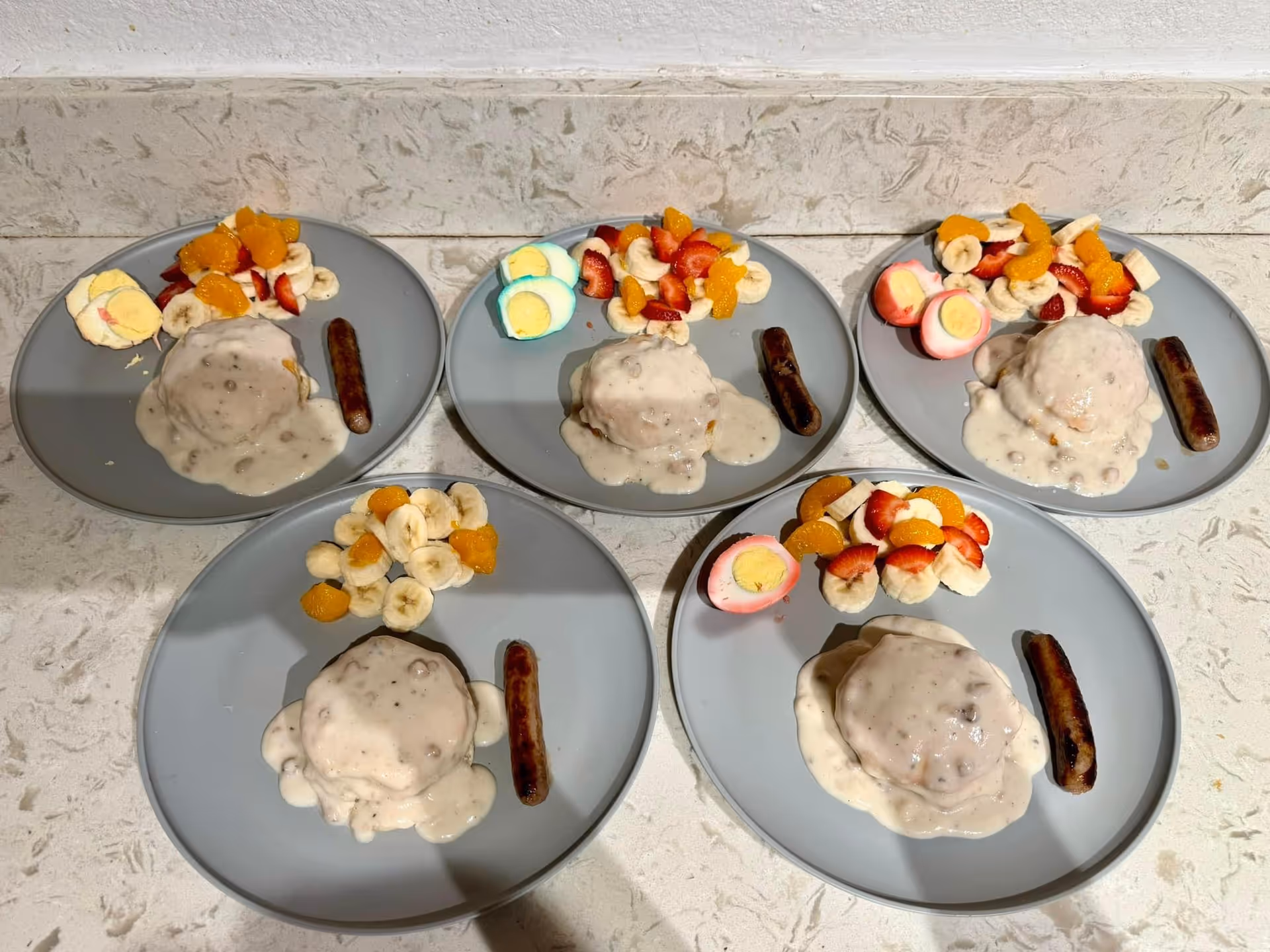 Five gray plates each with a serving of biscuits and gravy, a sausage link, and a side of sliced bananas, mandarin orange segments, strawberries, and a halved colored hard-boiled egg on a light-colored countertop.