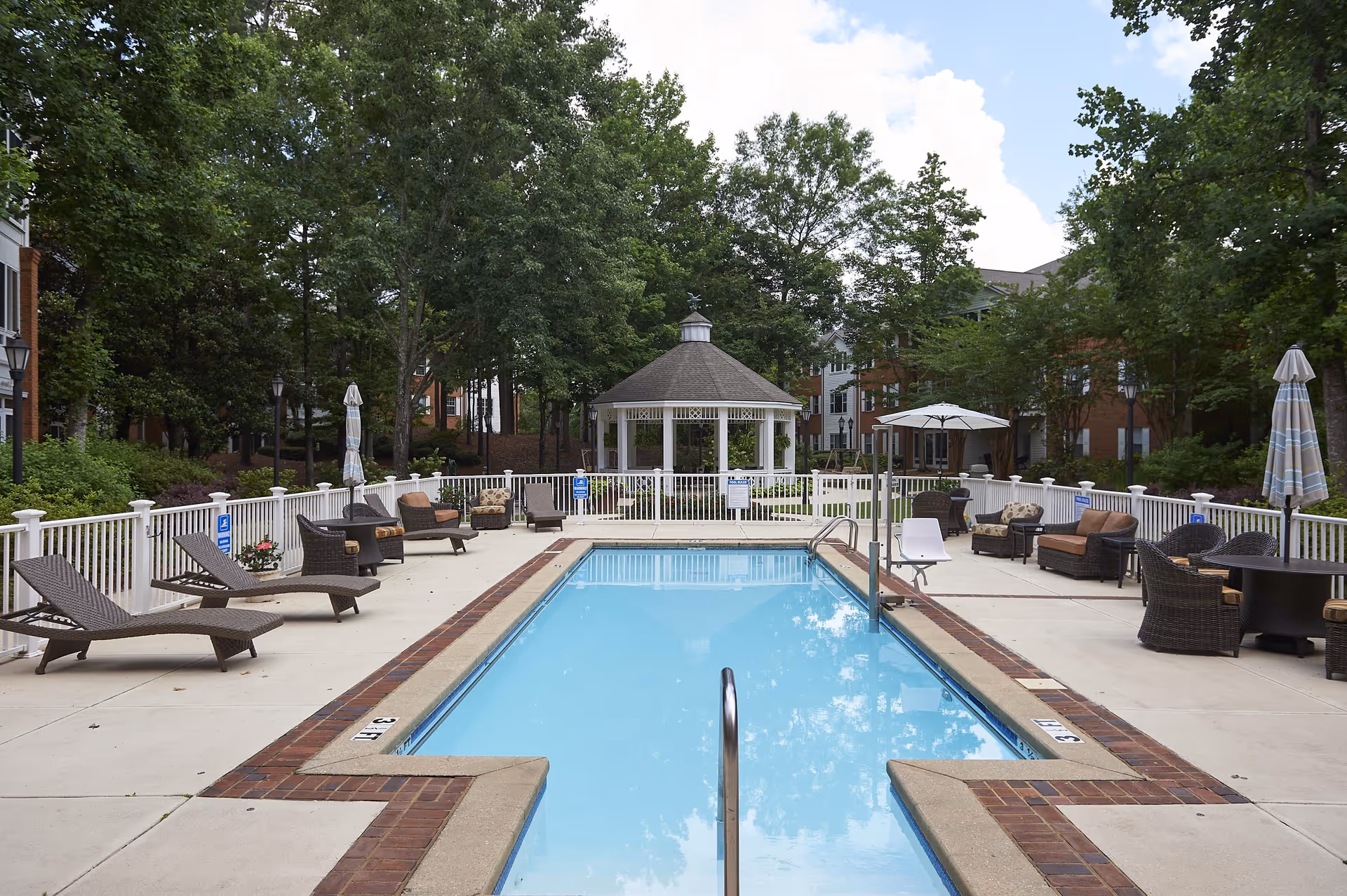 Outdoor swimming pool with lounge chairs, umbrellas, and a gazebo surrounded by trees and buildings.
