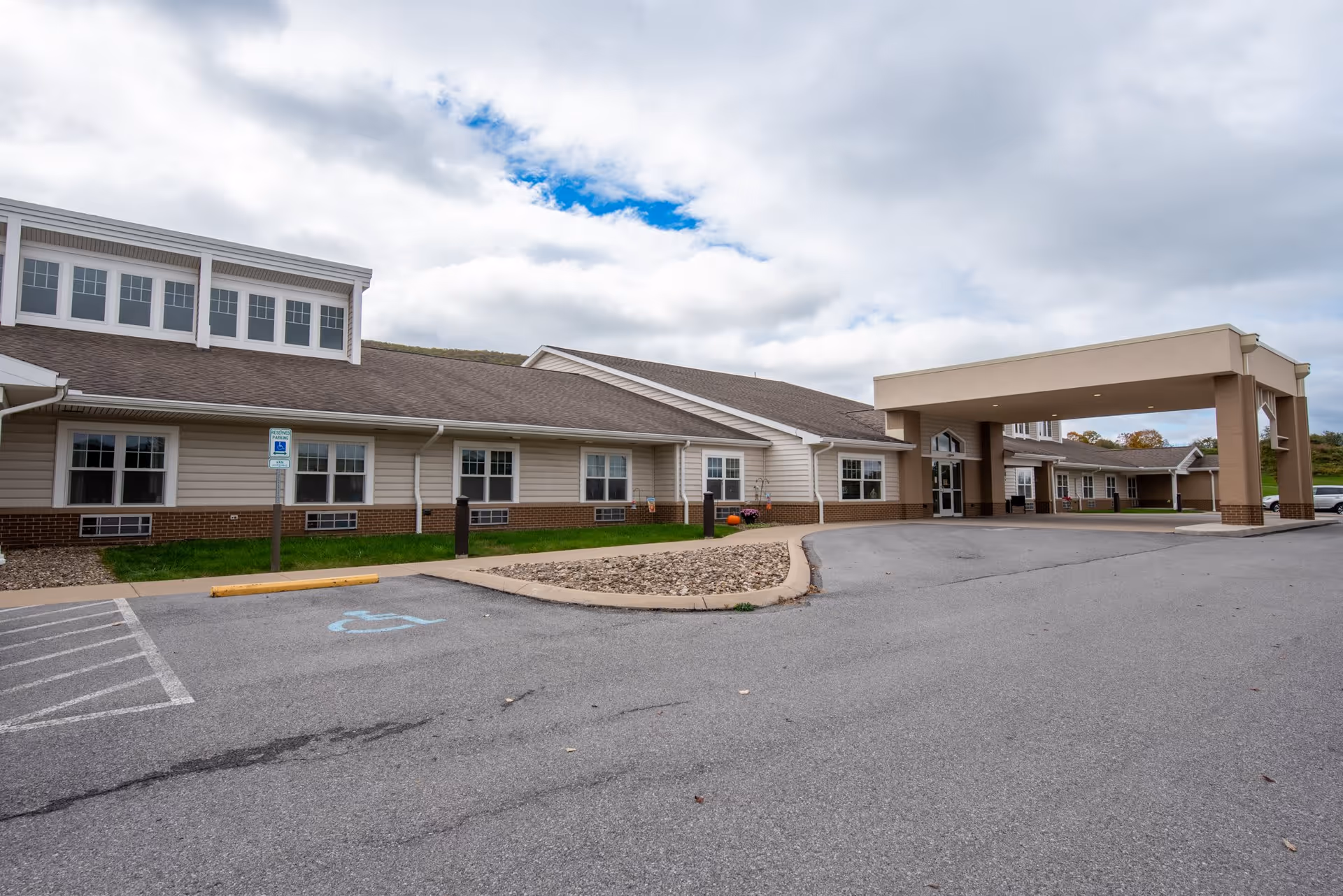 Front exterior of a single-story senior living facility with a covered entrance, windows, and a parking lot with a wheelchair space under a cloudy sky.