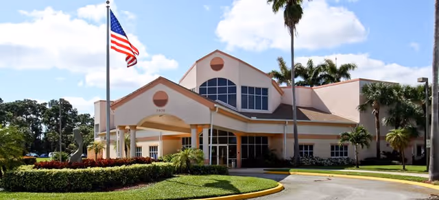 Exterior view of a senior living facility building with a covered entrance, large windows, palm trees, and an American flag on a flagpole in front. The sky is partly cloudy.