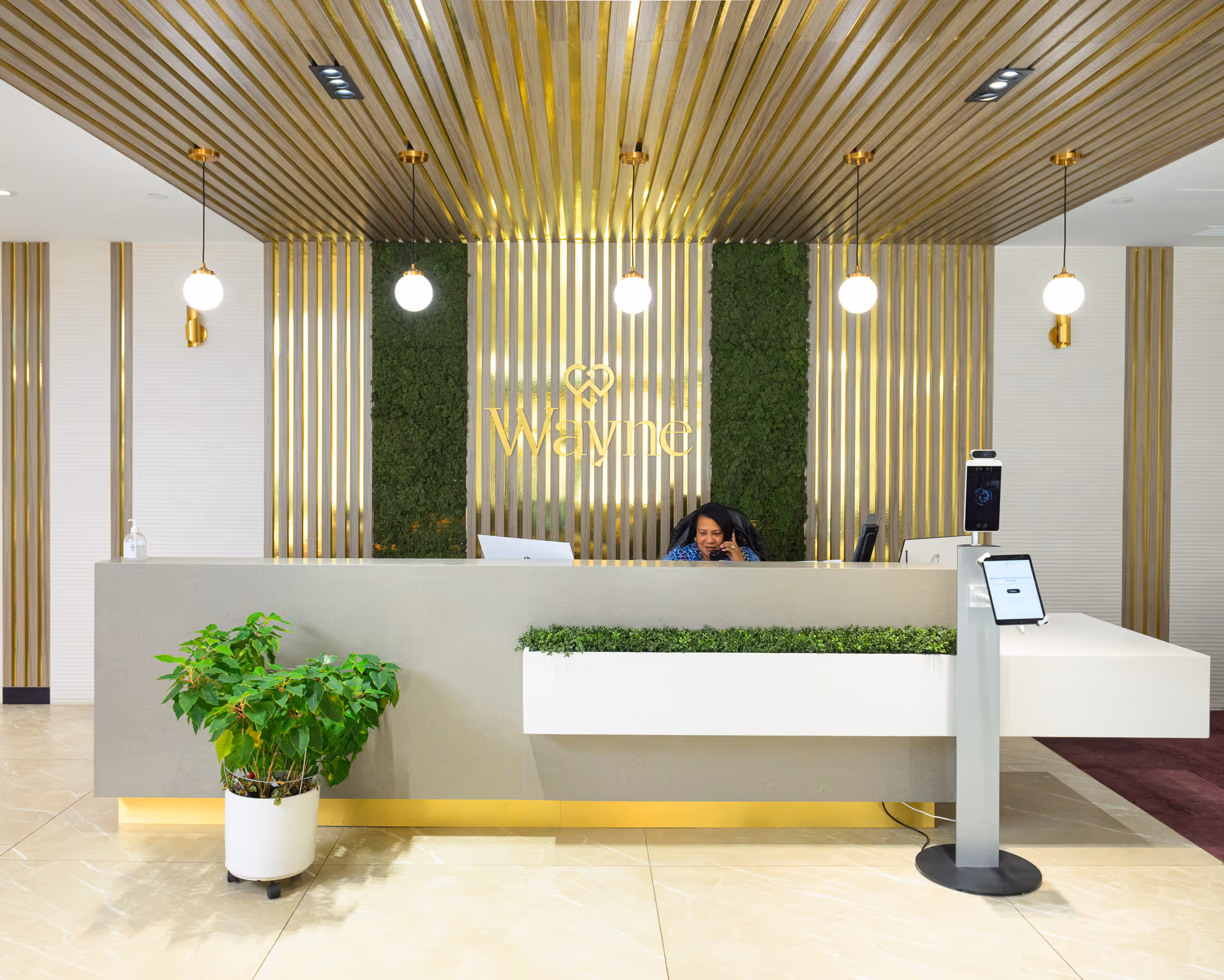 Reception area of The Wayne Center for Nursing and Rehabilitation featuring a modern front desk with a receptionist on the phone, decorative green plants, gold vertical accents on the wall, and hanging globe pendant lights.