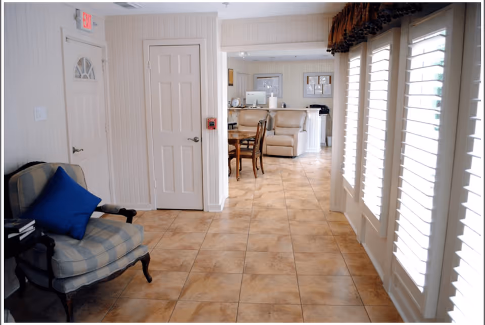 Interior view of a living area in an assisted living facility featuring a tiled floor, a cushioned armchair with a blue pillow on the left, a dining table with chairs in the middle, and two beige recliners near a counter in the background. Large windows with white shutters are on the right side, allowing natural light to fill the space.