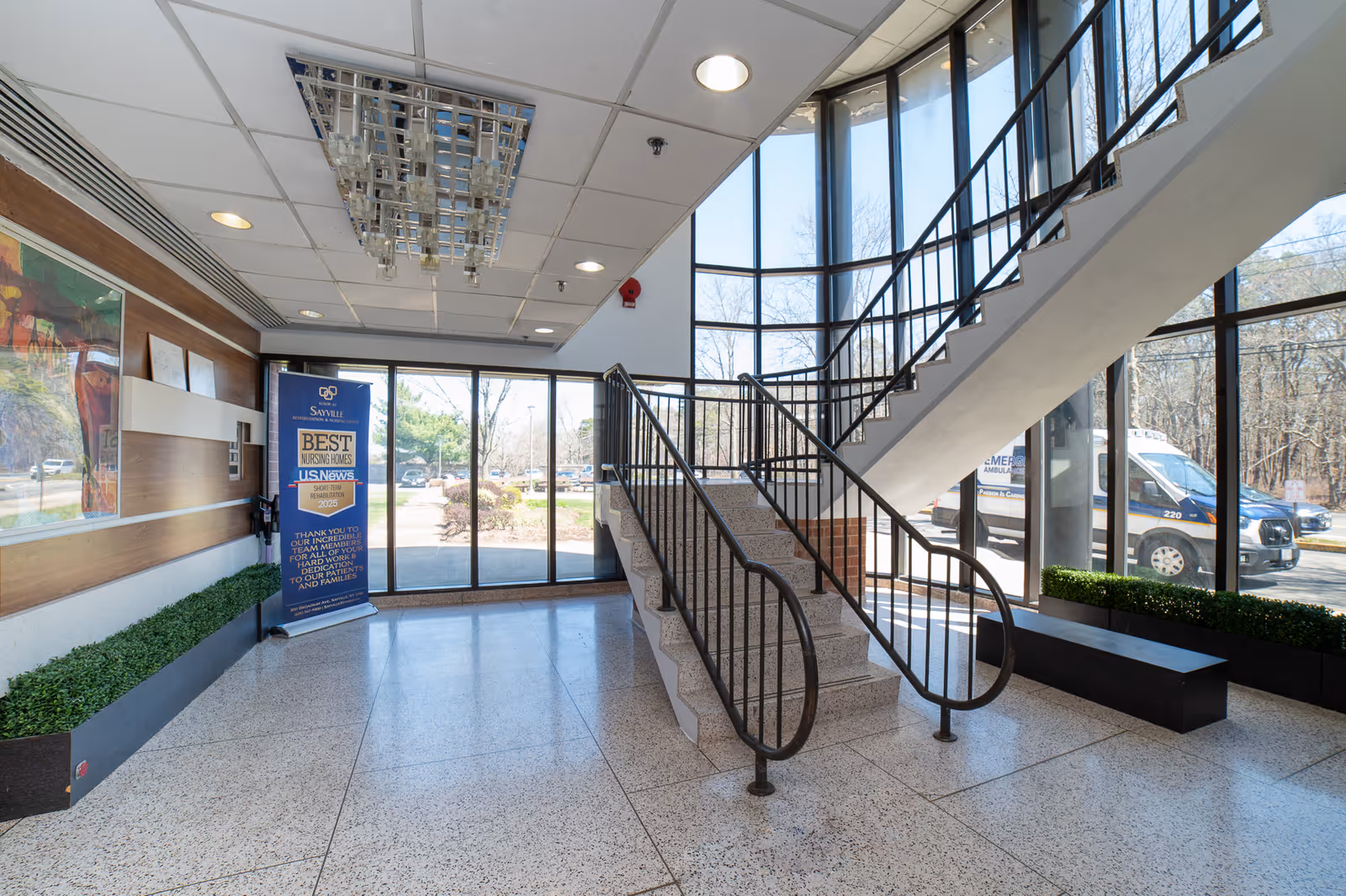 Interior view of a facility lobby with a staircase featuring black railings leading to an upper floor. Large floor-to-ceiling windows allow natural light to fill the space, showing a parking lot and trees outside. There is a blue banner near the entrance highlighting an award for Best Nursing Homes by U.S. News. The floor is terrazzo, and there are planters with green plants along the walls.