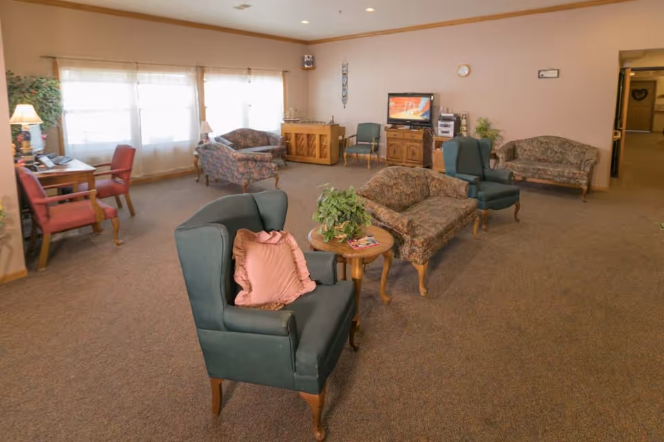 A cozy assisted living facility common area with multiple upholstered chairs and sofas arranged around small wooden tables. There is a television on a wooden cabinet against the far wall, a piano, and a desk with a lamp and chairs near large windows letting in natural light. The room has beige walls and carpeted floors.
