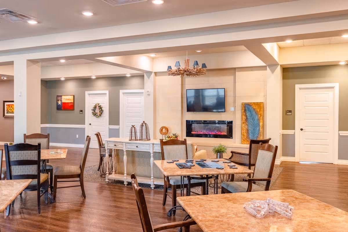 A well-lit common area with several tables and chairs arranged for socializing or activities. The room features a mounted flat-screen TV above a modern electric fireplace, decorative artwork on the walls, and a chandelier with blue lampshades. The flooring is wood, and there are white doors and trim with soft green and beige walls.