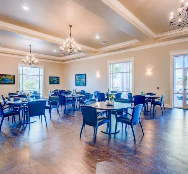 Bright communal dining room with round tables surrounded by blue chairs, chandeliers, and large windows.