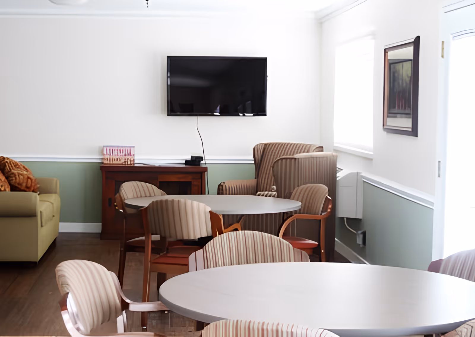 Bright common room with round tables and striped chairs, a wall-mounted TV, and a sofa.
