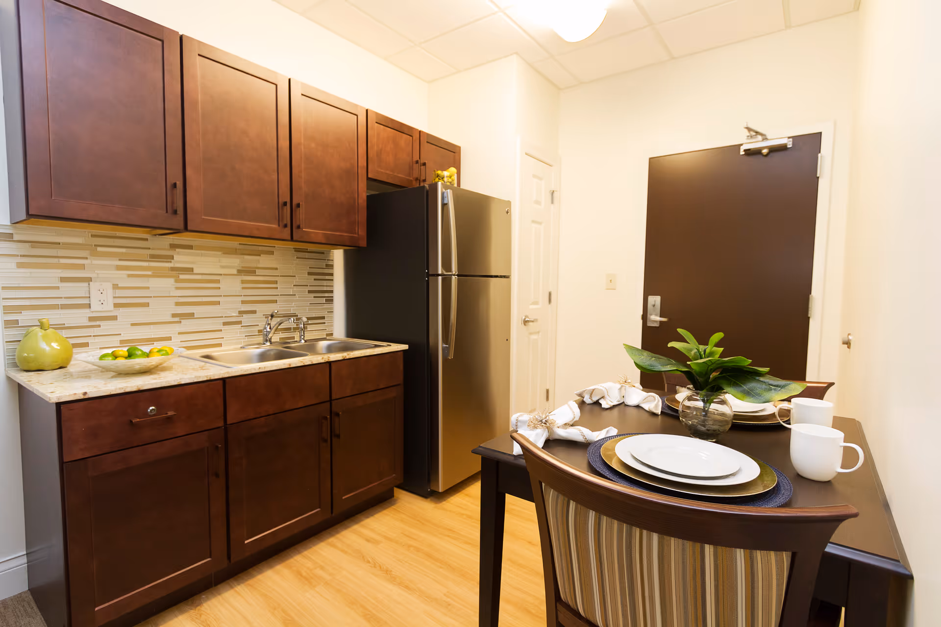 A small kitchen area with dark wooden cabinets, a stainless steel refrigerator, a double sink, and a tiled backsplash. To the right, there is a dining table set for two with plates, cups, napkins, and a small plant centerpiece. The floor is light wood, and there is a dark brown door in the background.