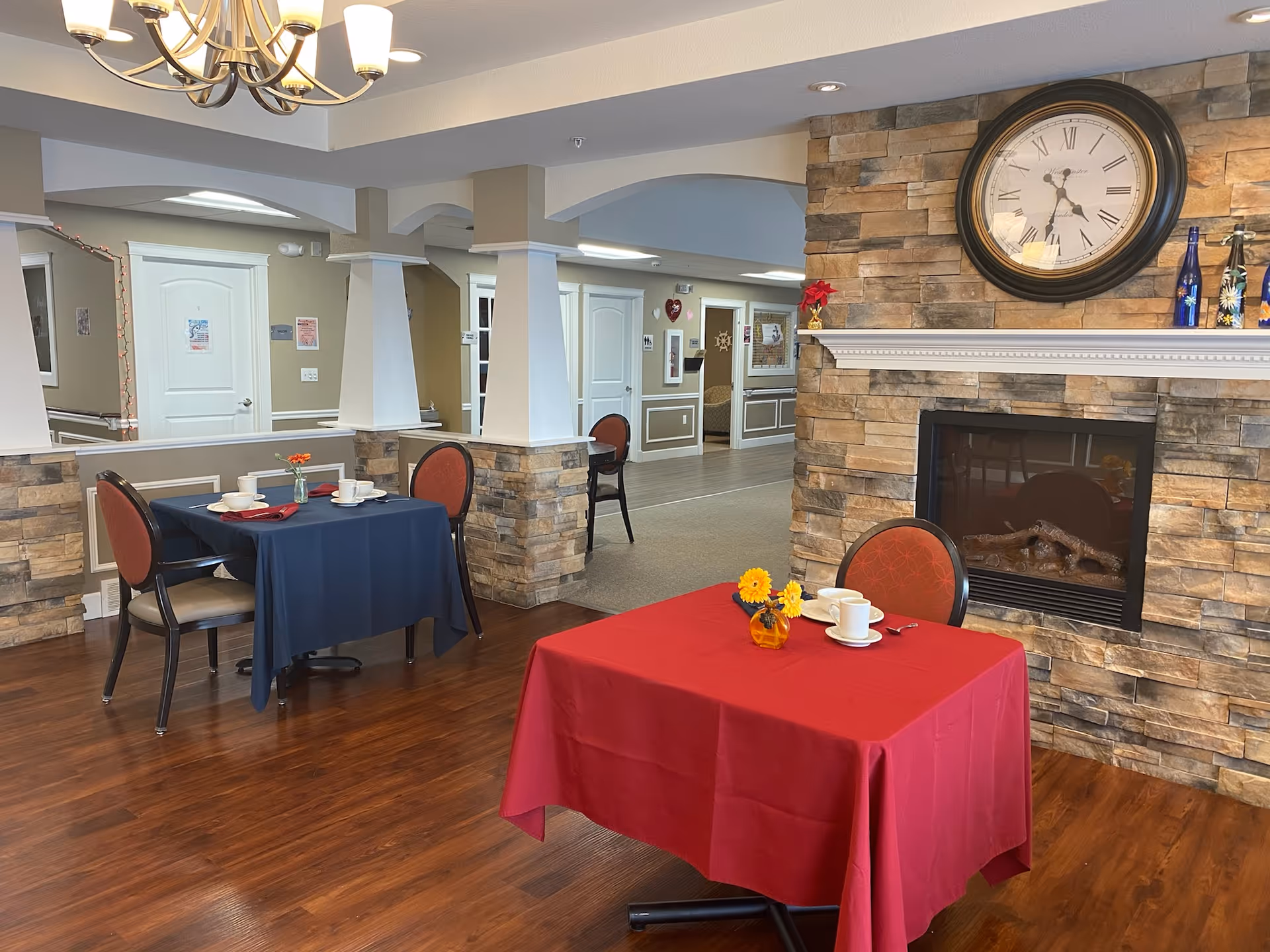 Interior dining area with two tables covered with red and blue tablecloths, each set with cups, saucers, and small flower vases. The room features a stone fireplace with a large clock above it, wooden flooring, and beige walls with white trim. There are chairs with red cushions around the tables and a chandelier hanging from the ceiling.