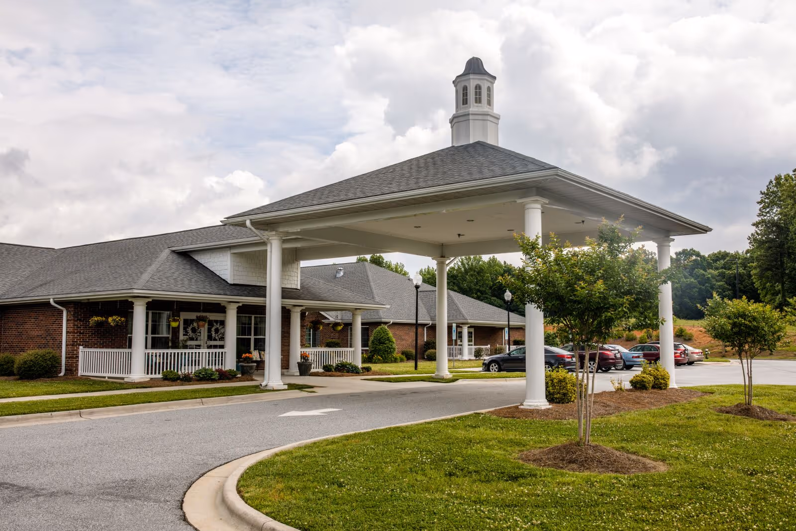 Exterior view of a single-story brick building with a large covered entrance supported by white columns. The building has a gray shingled roof with a small cupola on top. There are several small trees and shrubs planted around the driveway and parking area, where multiple cars are parked. The sky is partly cloudy.