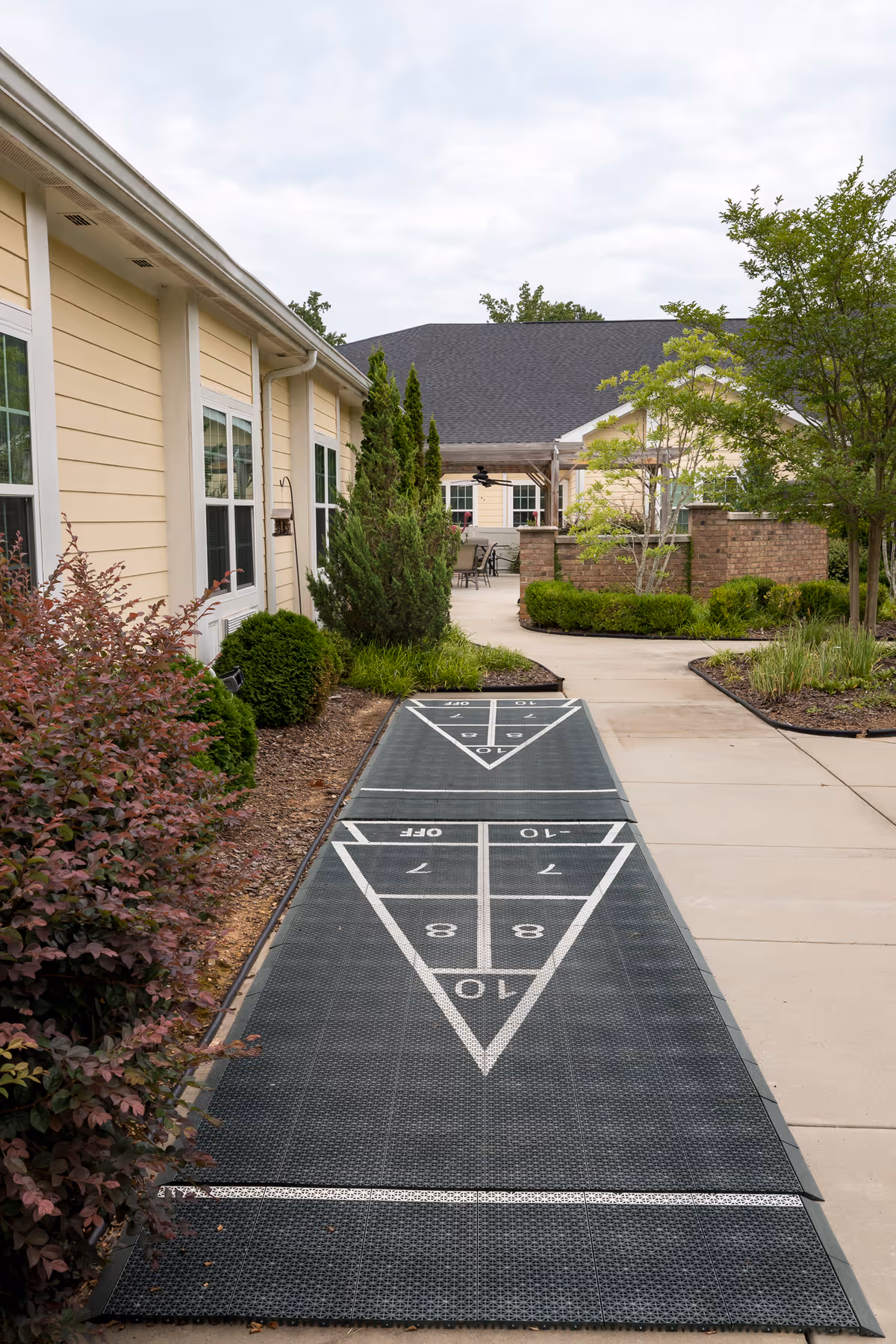 Outdoor shuffleboard court with a pathway leading to a patio area surrounded by bushes, trees, and a yellow building with white trim.