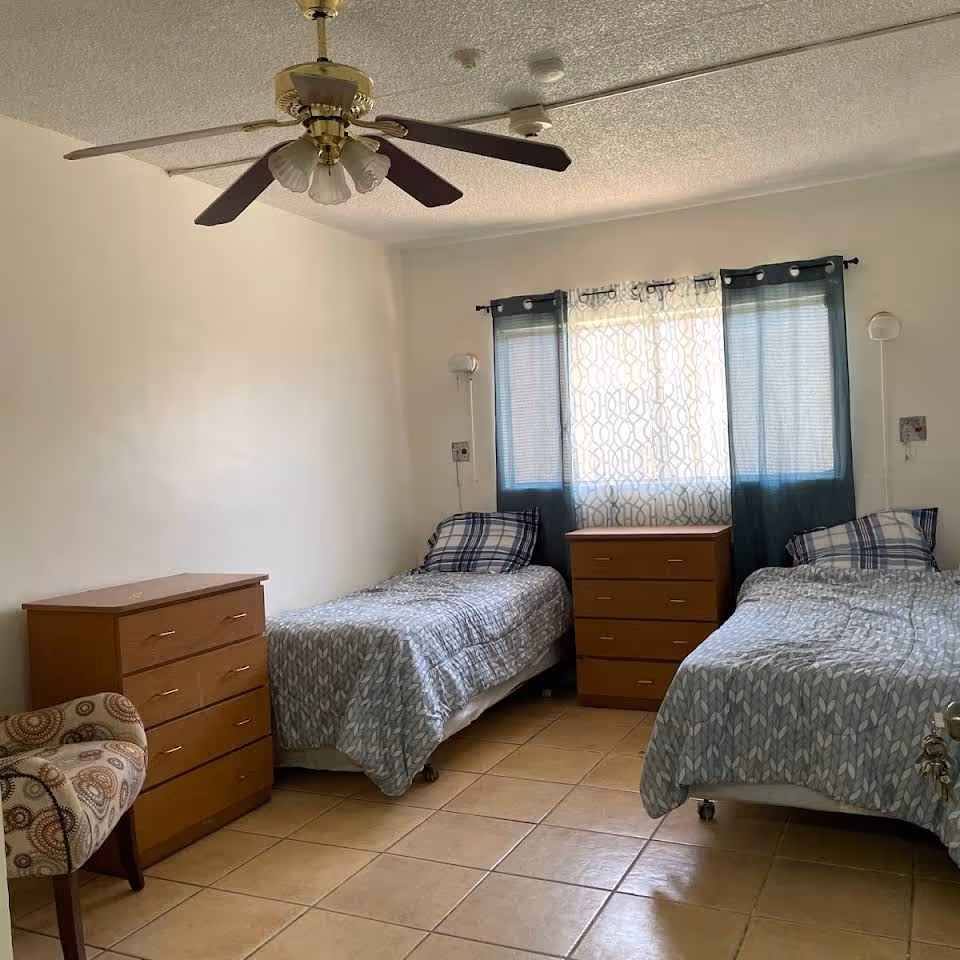 A bedroom with two single beds, each with a gray patterned bedspread and a plaid pillow. Between the beds is a wooden dresser under a window with blue curtains and a sheer patterned curtain. There is a wooden chest of drawers on the left side of the room and a patterned upholstered chair in the corner. The ceiling has a fan with five blades and light fixtures.