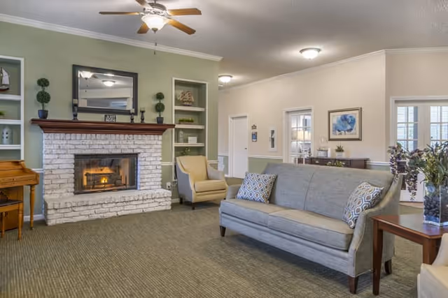 A cozy living room area with a white brick fireplace, a ceiling fan with light, a gray sofa with patterned pillows, beige armchairs, built-in shelves with decorative items, a wooden piano, and framed artwork on the walls.
