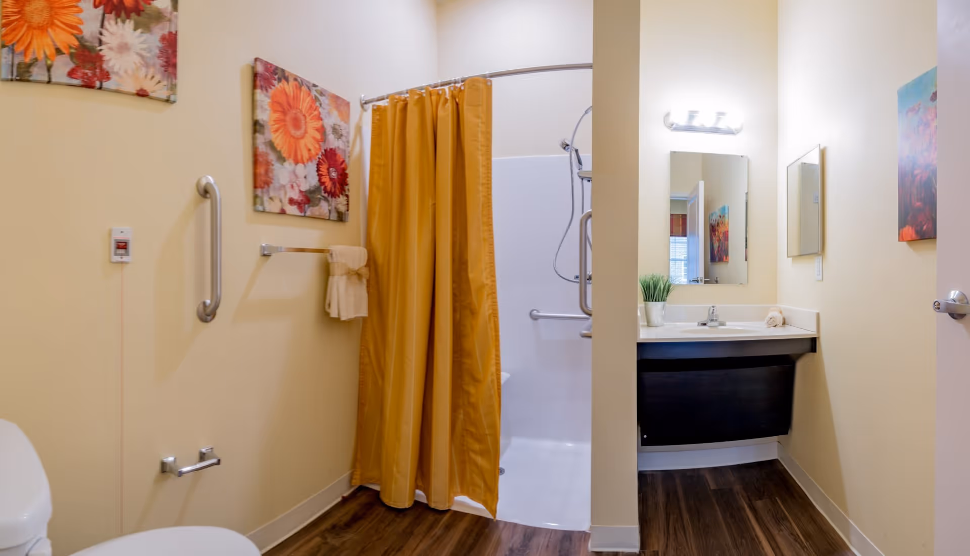 A bathroom with a white toilet, a shower with a yellow curtain, a sink with a mirror above it, and floral artwork on the walls. The floor is wood-style, and there are grab bars installed near the toilet and in the shower for accessibility.