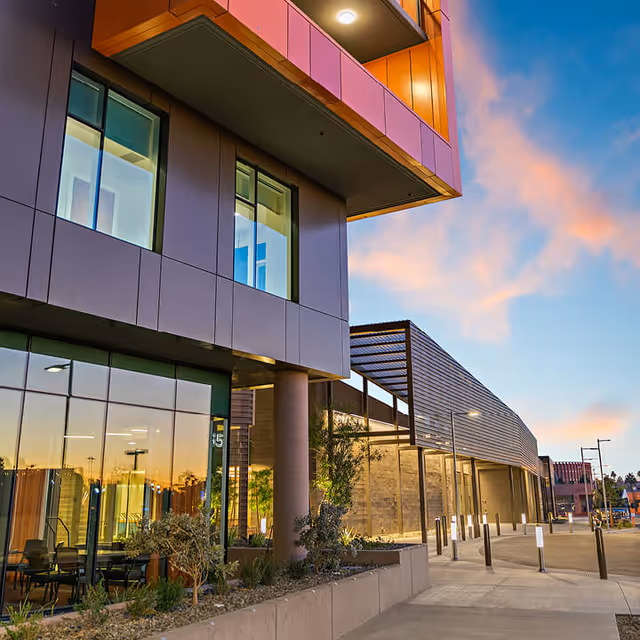 Modern building exterior at sunset with large windows, a covered walkway, and landscaped plants along the sidewalk under a partly cloudy sky.