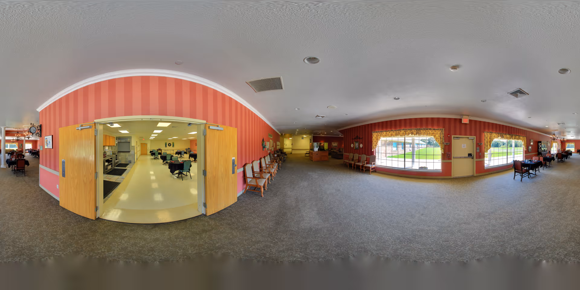 Wide interior view of a senior living facility common area with open double doors to a dining room, rows of chairs, tables and large windows overlooking a lawn.