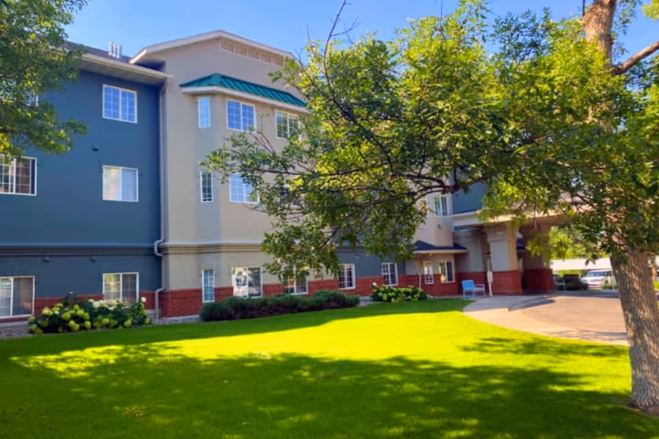 Exterior view of a senior living facility building with a well-maintained green lawn and trees in the foreground. The building has multiple windows, a covered entrance, and a mix of beige, blue, and red brick exterior walls under a clear sky.