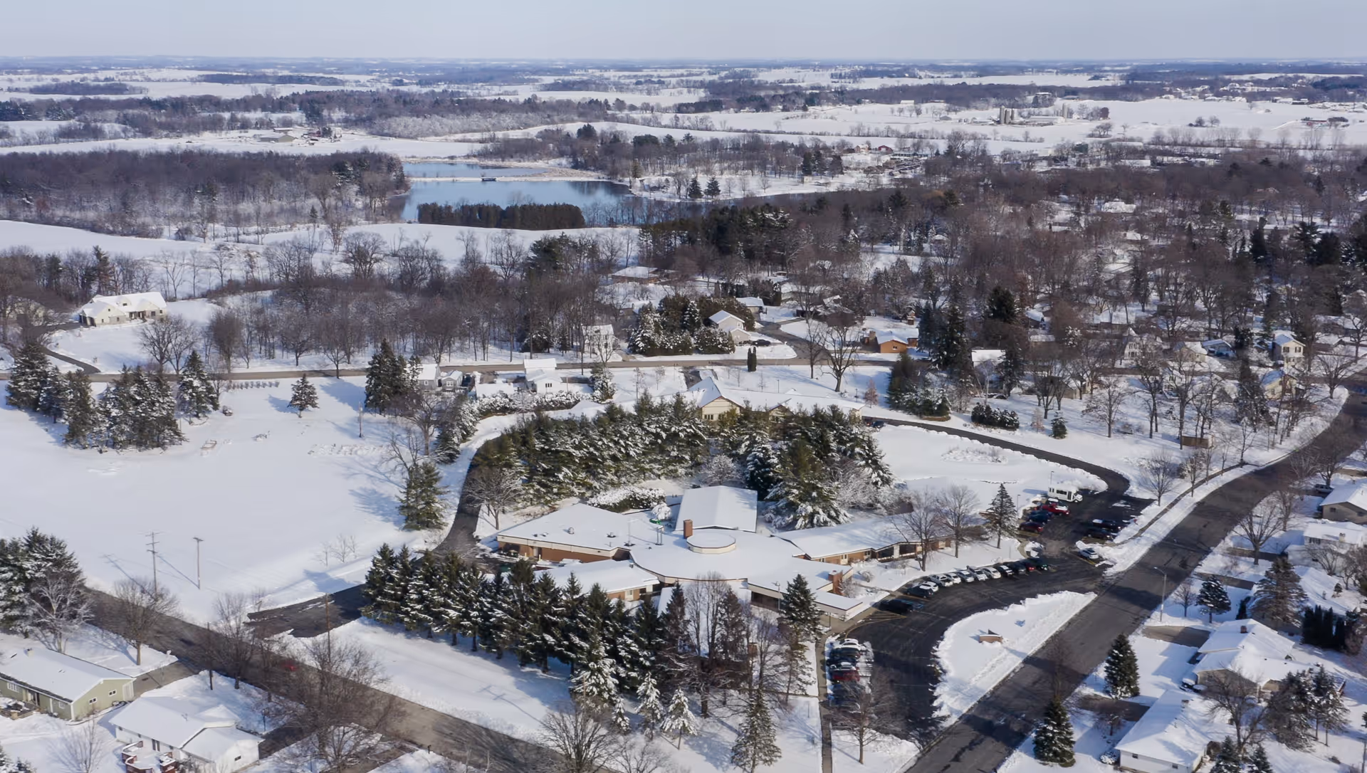 Aerial view of Evansville Manor Skilled Nursing facility and surrounding snowy landscape, including roads, trees, houses, and a partially frozen lake in the distance.