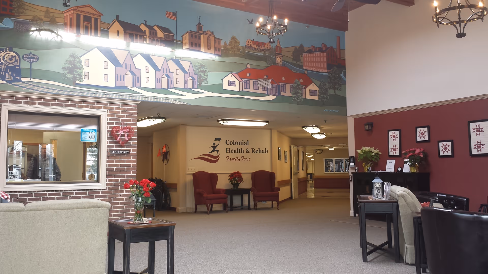 Interior view of the Colonial Health & Rehab Center of Plainfield, LLC lobby area featuring a reception window on the left, a mural of a townscape above, two red armchairs with a small table and flowers in the center, and a seating area with chairs and tables on the right. The walls are decorated with framed artwork and plants, and chandeliers hang from the ceiling.