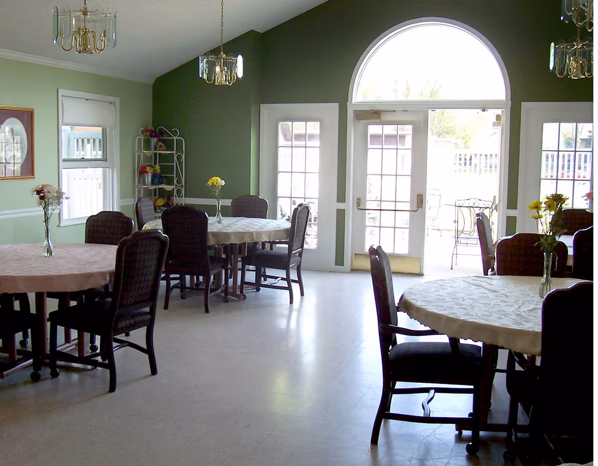 A dining room with round tables covered in light-colored tablecloths, each table having a vase with flowers. Several dark cushioned chairs surround the tables. The room has green walls, large windows, and a glass door with an arched window above it letting in natural light. Two chandeliers hang from the ceiling.