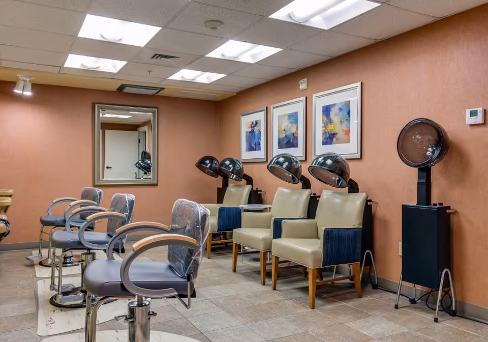 Interior view of a salon area with several salon chairs and hair drying stations against a peach-colored wall. There are framed abstract paintings on the wall and a large mirror reflecting part of the room. The floor is tiled and the ceiling has fluorescent lighting.