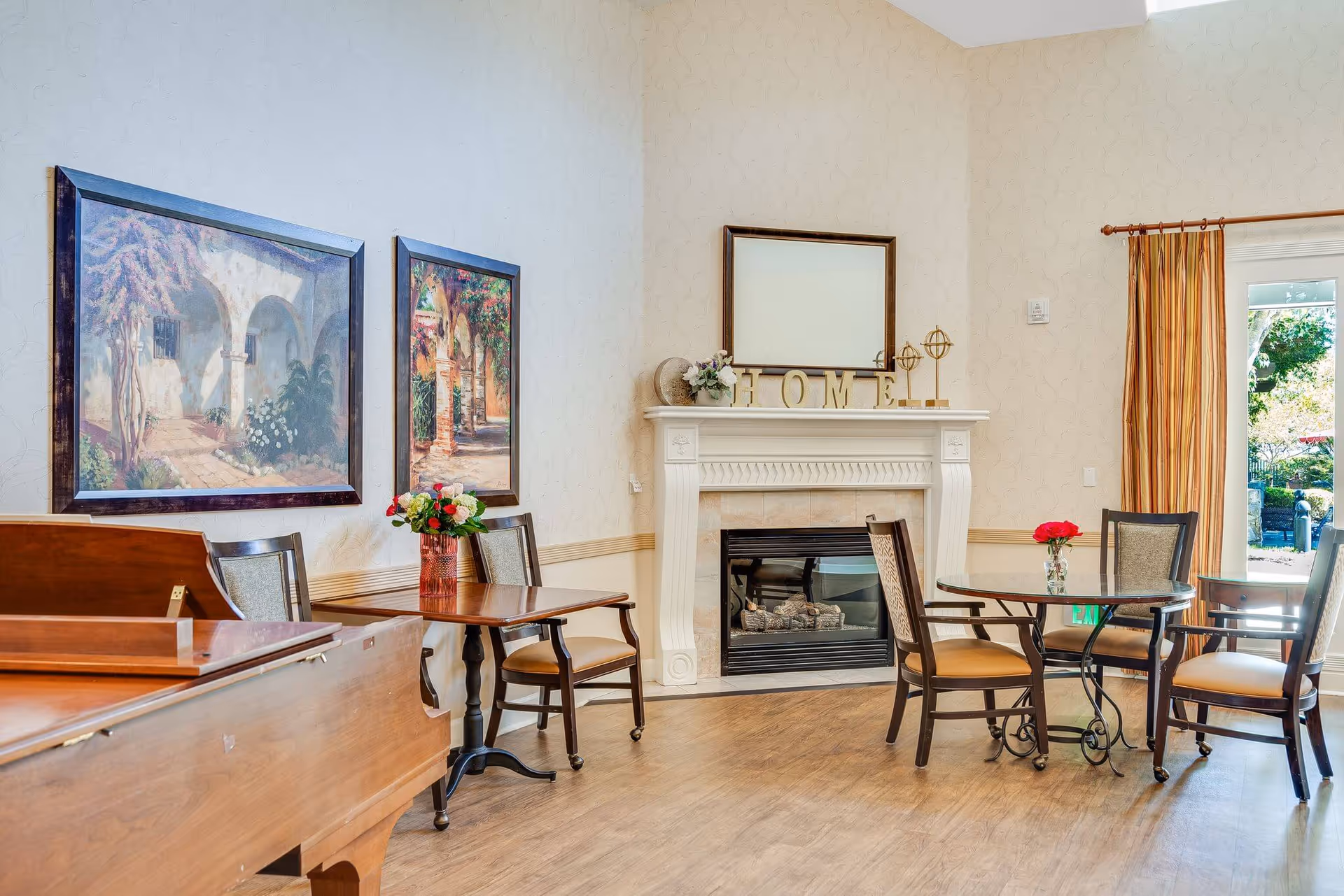 Bright common room with a fireplace, piano, tables and chairs, framed wall art, and a doorway to the outdoors.