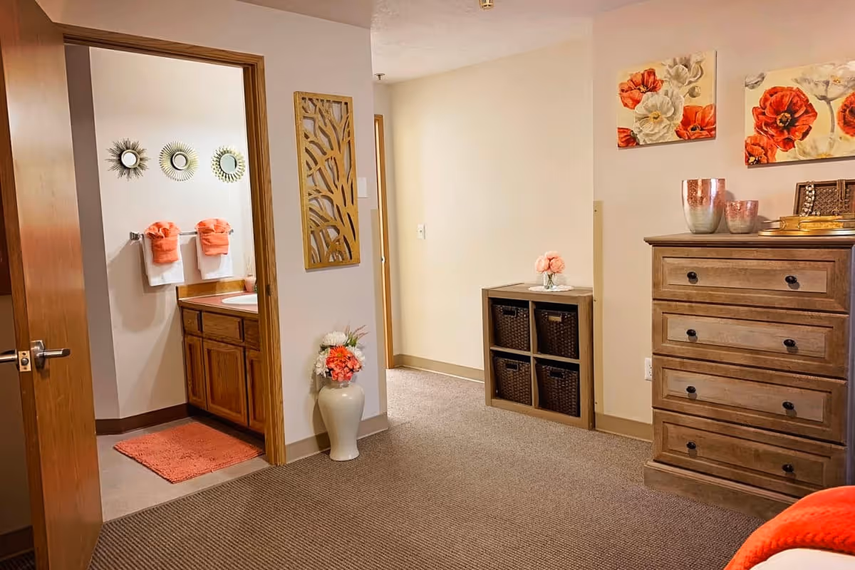Interior view of a senior living facility room showing a wooden dresser with decorative items and floral paintings above it. To the left, there is an open door leading to a bathroom with a sink, mirror, and orange towels. The room has beige walls, carpeted floor, and a small storage unit with baskets and a flower vase on top.