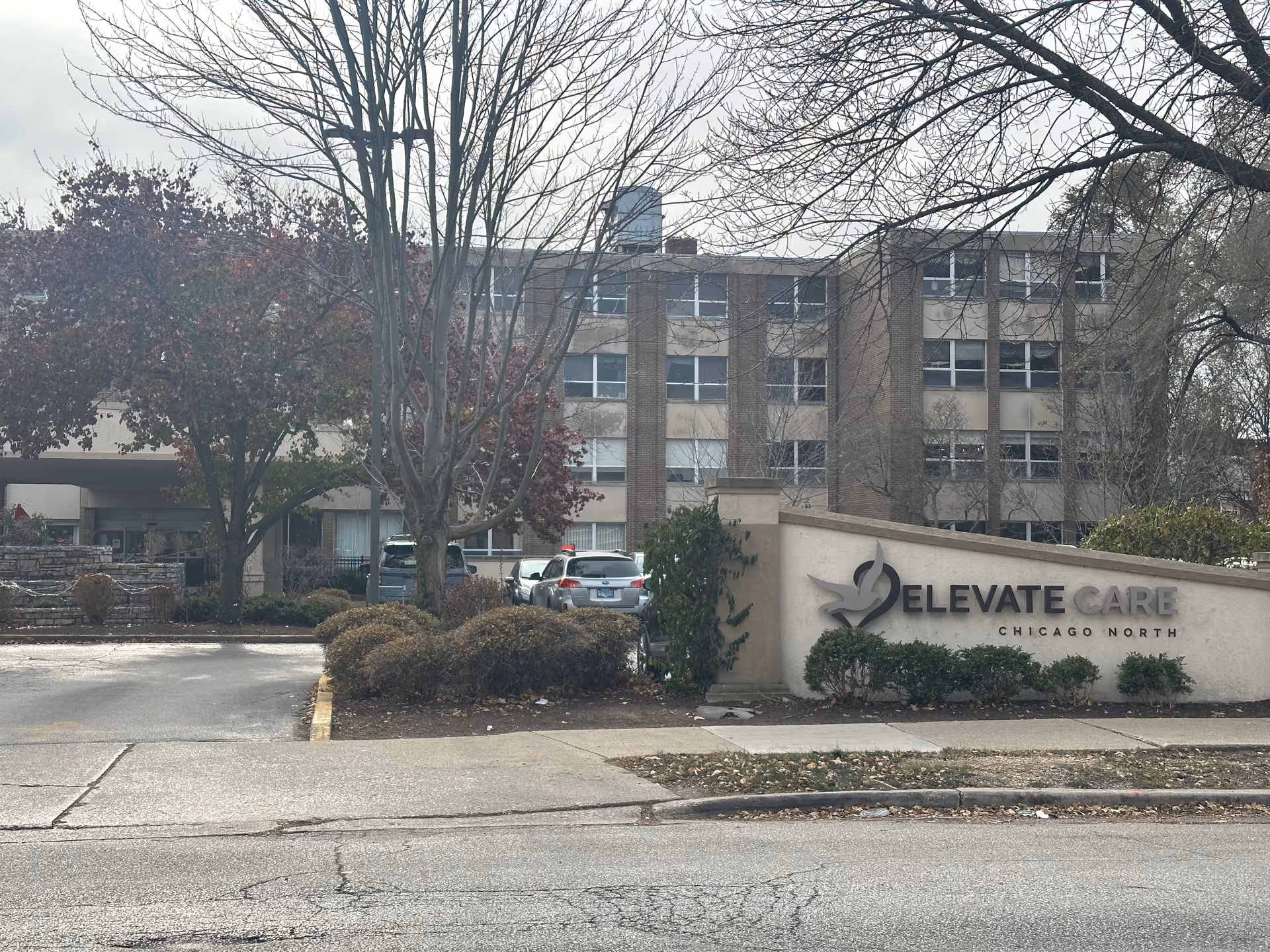 Exterior view of a multi-story brick building with several windows, surrounded by leafless trees and bushes. In front of the building is a large sign that reads 'Elevate Care Chicago North'. Several cars are parked near the entrance.
