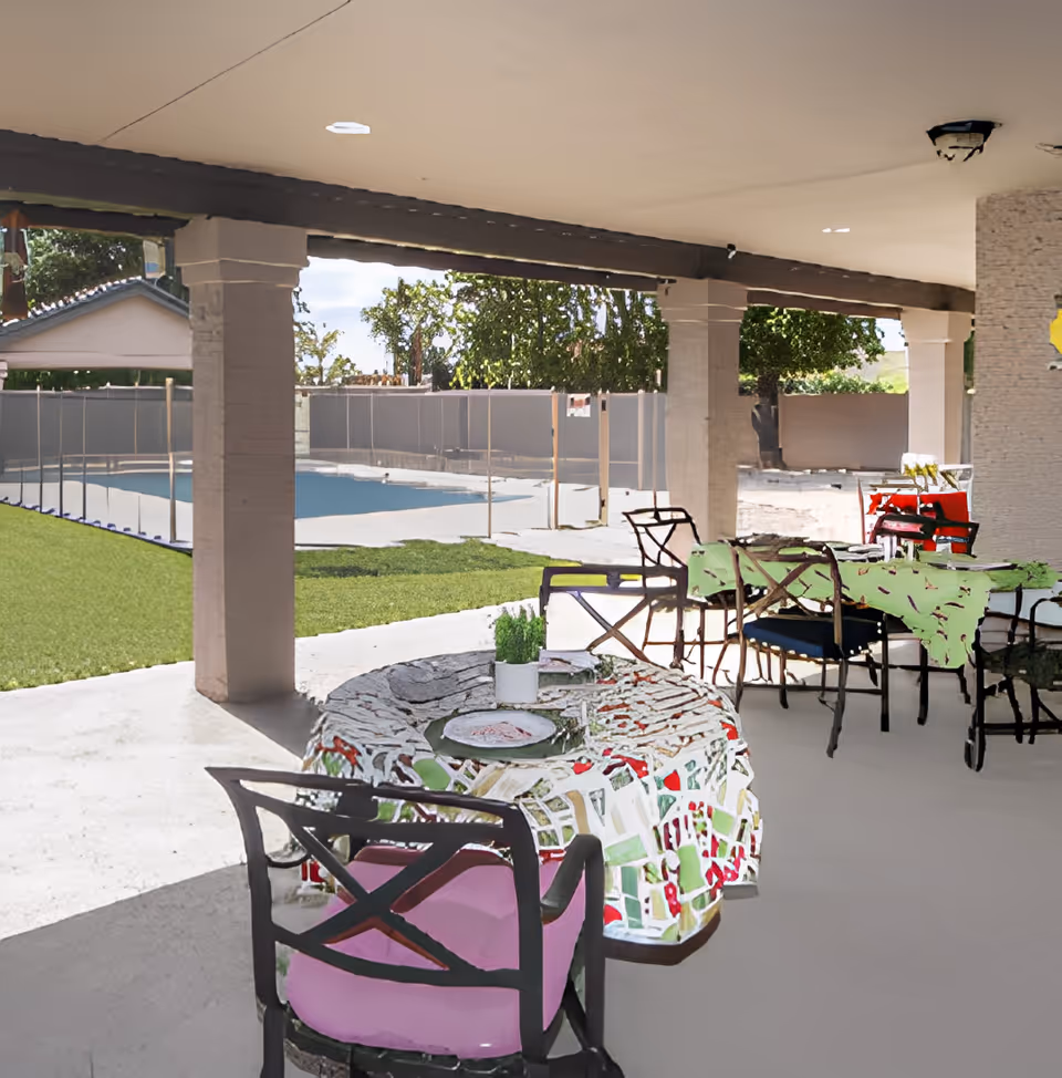 Covered outdoor patio with tables and chairs overlooking a fenced swimming pool and lawn.